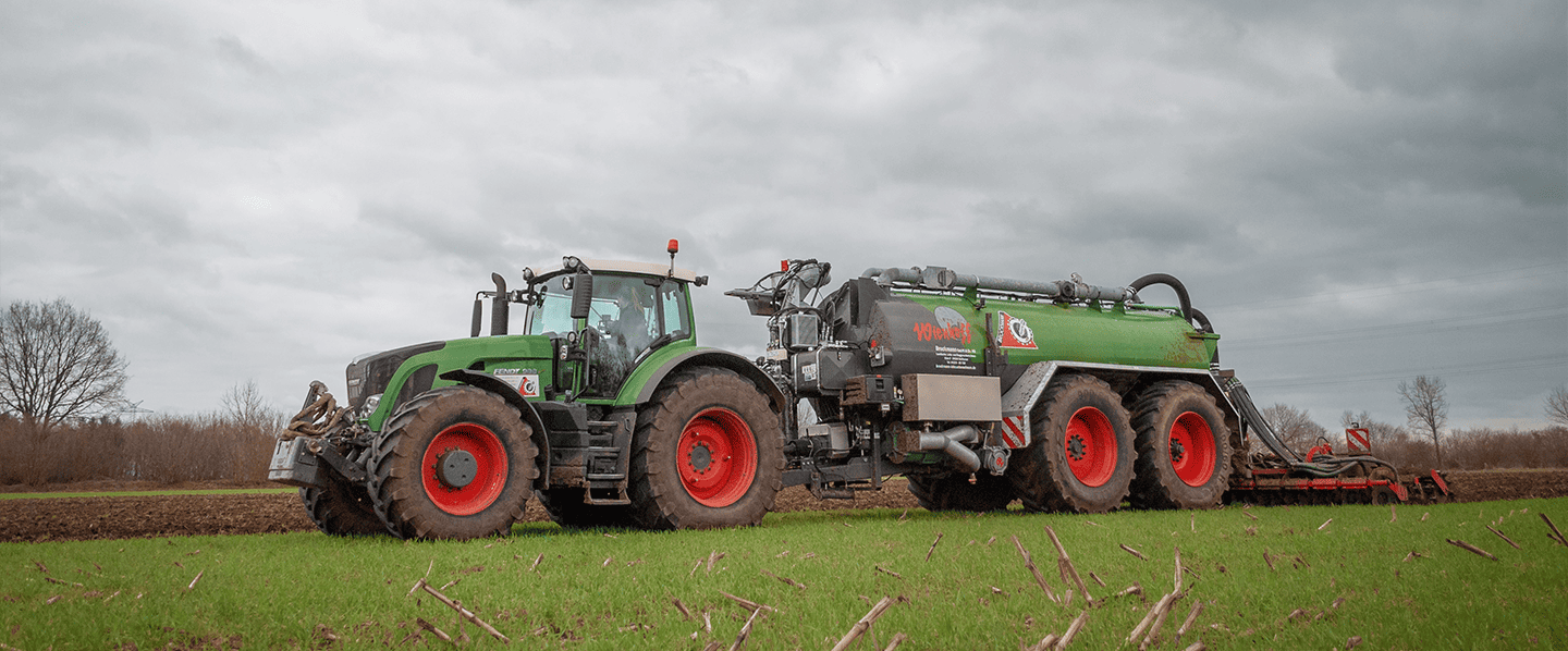 Ein Fendt-Traktor zieht einen grünen Gülle-Anhänger auf einem Feld unter bewölktem Himmel in Schleswig-Holstein.